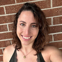 A photo of Kathryn Jaksic. She has wavy brown hair and is wearing a dark top and a gold pendant necklace, standing in front of a red brick wall in this photo.