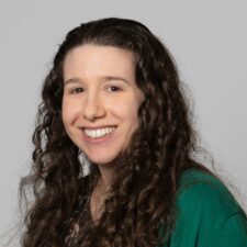 Photo of Keri Lemoine. Shows a woman with long, dark brown, tightly curled hair that falls past her shoulders. She is wearing a green cardigan over a black blouse with a colorful leaf-like pattern. The background is plain light gray.
