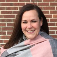 Person with long, straight dark brown hair standing in front of a red brick wall, wrapped in a large scarf.