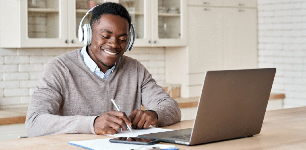 A smiling young man wearing headphones, sitting in front of a laptop. He's writing on paper in front of the laptop.