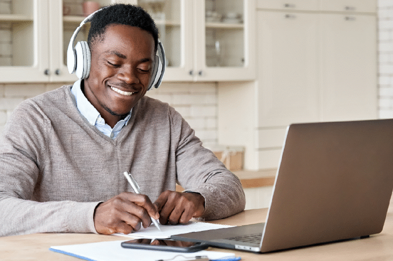 A smiling young man wearing headphones, sitting in front of a laptop. He's writing on paper in front of the laptop.