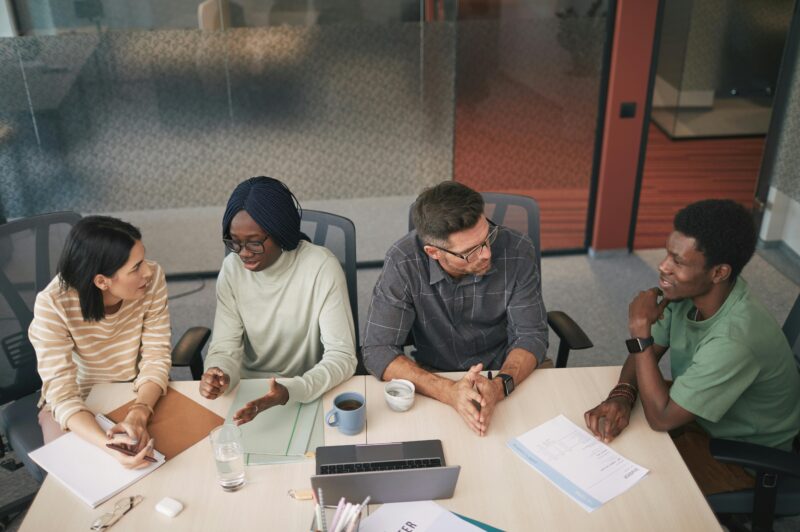 A group of four people sit around a conference table in a modern office, engaged in discussion. They are seated in a row and facing one another, with notebooks, papers, a laptop, and drinks spread across the table. A glass wall and office furniture are visible in the background.